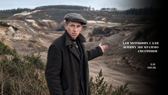 Уцілілий в'язень Сирецького концтабору Юхим Вілкіс дає свідчення та вказує на місце масових страт в урочищі Бабин Яр. Кадр із документальної кінохроніки «Табір військовополонених у Дарниці; Бабин Яр» («The POW camp in Darnytsia; Babyn Yar»), листопад 1943 року. Чорно-білий скріншот із документальної кінохроніки з вертикальними лініями перешкод плівки. По центру великим планом зафіксовано Юхима Вілкіса — уцілілого свідка нацистських злочинів. Він одягнений у темне довге пальто та кепку, його обличчя напружене, а ліва рука піднята у жесті, що вказує назад на панораму глибокого й розлогого Бабиного Яру. На передньому плані праворуч внизу видно металеву деталь кінематографічного або військового обладнання.